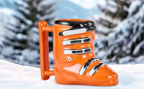 ceramic ski boot mug in orange, holds 14 ounces and is sitting on a snowy pine tree background.